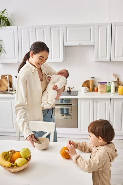 Mulher atraente alegre preparando café da manhã para seu filho criança enquanto segurando seu menino recém-nascido — Fotografia de Stock
