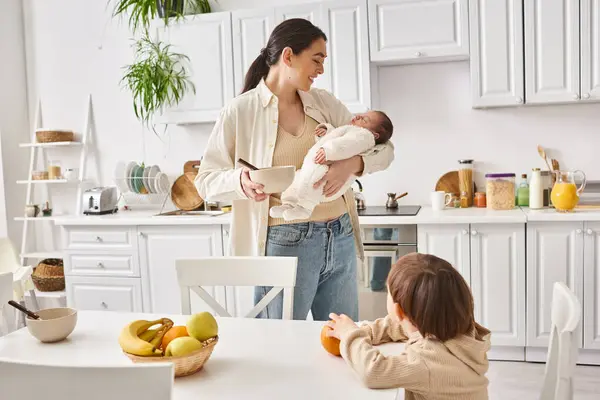 Joyful attractive woman preparing breakfast for her toddler son while holding her newborn baby boy — Stock Photo