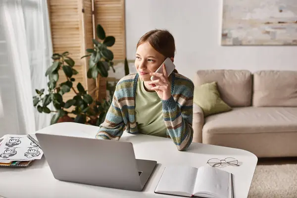 Sonriente adolescente haciendo una llamada telefónica mientras está sentado cerca de la computadora portátil en el escritorio, sesión de estudio electrónico - foto de stock