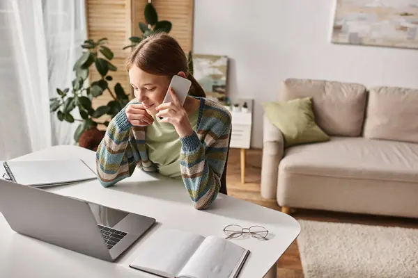 Alegre adolescente chica haciendo una llamada telefónica mientras está sentado cerca de la computadora portátil en el escritorio, sesión de estudio electrónico - foto de stock