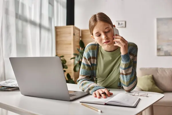 Chica adolescente enfocada haciendo una llamada telefónica mientras está sentada cerca de la computadora portátil en el escritorio, sesión de estudio electrónico - foto de stock