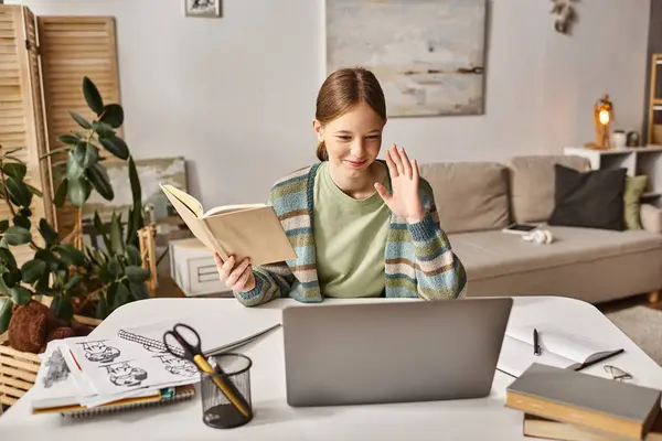 Happy gen z girl holding book while using a laptop during video call at home, teen lifestyle concept — Stock Photo