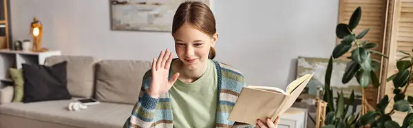 Horizontales Banner eines glücklichen Teenie-Mädchens, das Buch in der Hand hält, während es zu Hause winkt, zu Hause studiert — Stockfoto