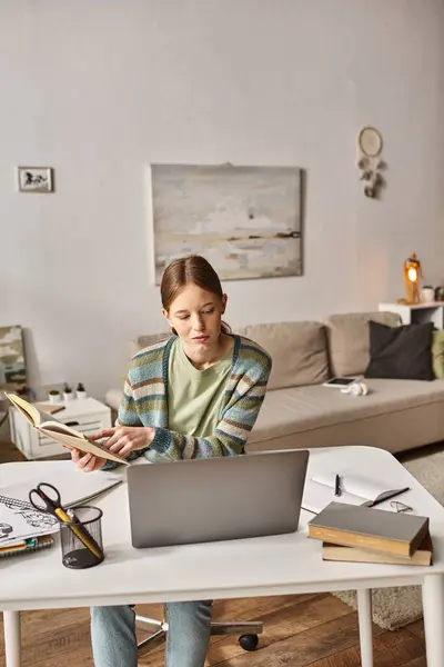 Adolescente sosteniendo libro durante el uso de un ordenador portátil durante la videollamada en casa, gen z concepto de estilo de vida - foto de stock