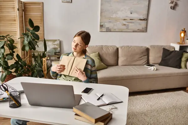 Adolescent fille tenant livre et en utilisant un ordinateur portable lors d'un appel vidéo à la maison, concept de style de vie gen z — Photo de stock