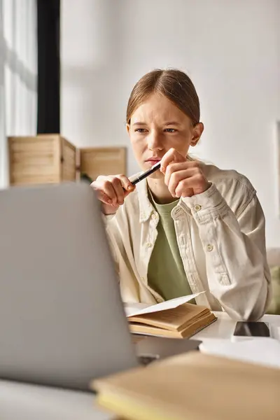 Adolescente sosteniendo la pluma y mirando a la computadora portátil mientras hace su tarea cerca del libro y el teléfono en el escritorio - foto de stock