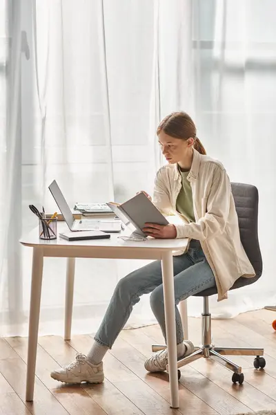 Teenager girl reading book while sitting near laptop and stationery on desk, online education — Stock Photo