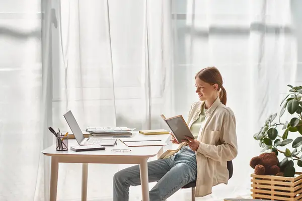 Glückliches Teenager-Mädchen liest Buch, während es neben Laptop und Schreibwaren auf dem Schreibtisch sitzt, Online-Bildung — Stockfoto