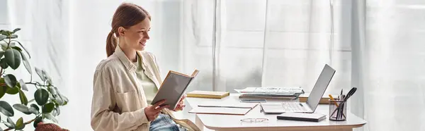 Livre de lecture adolescent heureux et assis près d'un ordinateur portable sur le bureau, éducation en ligne, bannière horizontale — Photo de stock