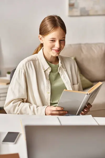 Fröhliches Teenager-Mädchen liest Buch, während es neben Laptop und Schreibwaren auf dem Schreibtisch sitzt, Online-Bildung — Stockfoto