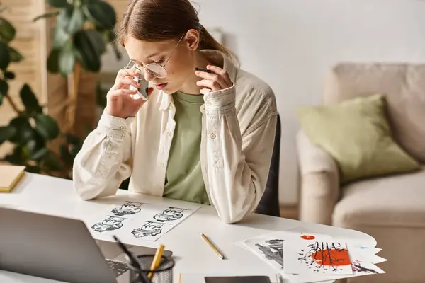 Дівчина-підліток розмовляє на смартфоні і дивиться на її ескіз малюнок біля ноутбука вдома — стокове фото