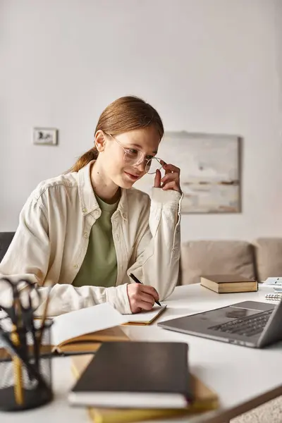 Teenage girl in glasses holding pen and writing during online class on laptop, taking notes — Stock Photo