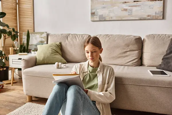 Chica adolescente pensativa leyendo su cuaderno cerca del sofá con auriculares y tableta digital cerca - foto de stock