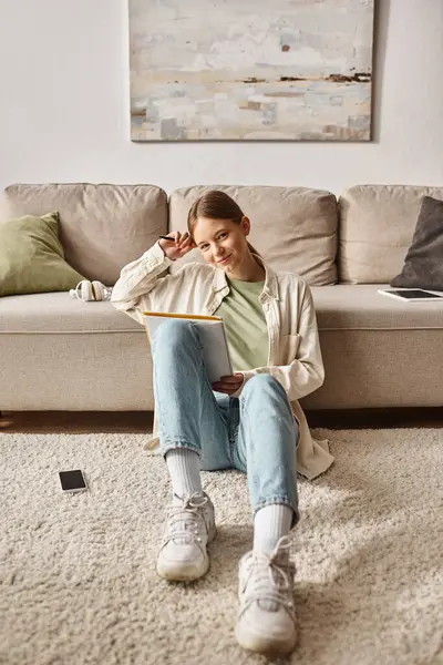 Chica adolescente pensativa leyendo su cuaderno y sentado cerca del sofá con teléfono inteligente cerca - foto de stock
