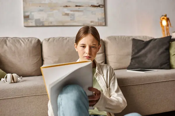 Chica adolescente pensativa leyendo su cuaderno cerca del sofá con auriculares y tableta digital - foto de stock