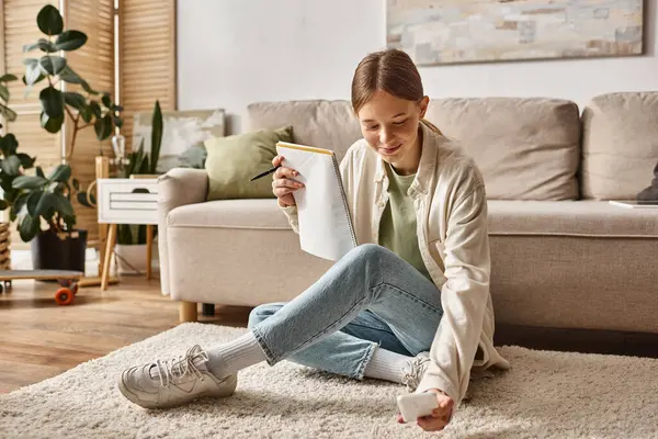 Feliz adolescente sosteniendo su portátil y teléfono inteligente y sentado cerca del sofá con auriculares - foto de stock