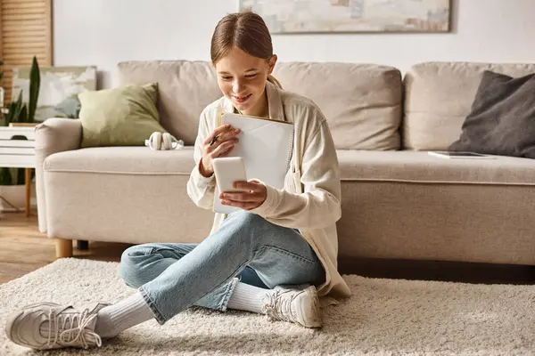 Adolescente feliz sosteniendo su portátil y teléfono inteligente y sentado cerca del sofá con auriculares - foto de stock