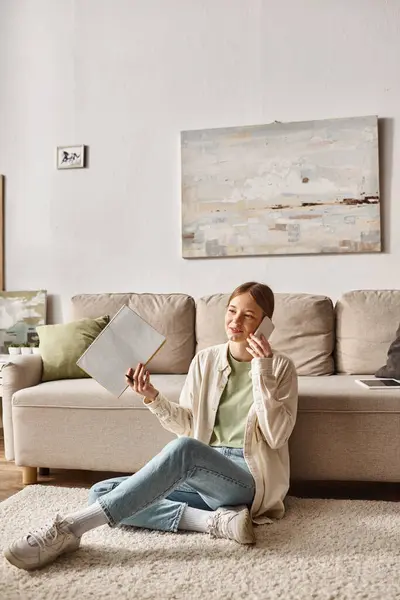 Happy teenager girl holding her notebook and talking on smartphone while sitting near the couch — Stock Photo