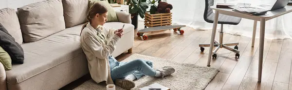 Young teenager girl using her smartphone while sitting on carpet near the couch in living room — Stock Photo