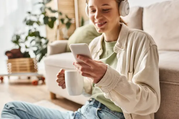 Menina adolescente sorridente em fones de ouvido sem fio usando smartphone enquanto segurando xícara de chá em casa — Fotografia de Stock
