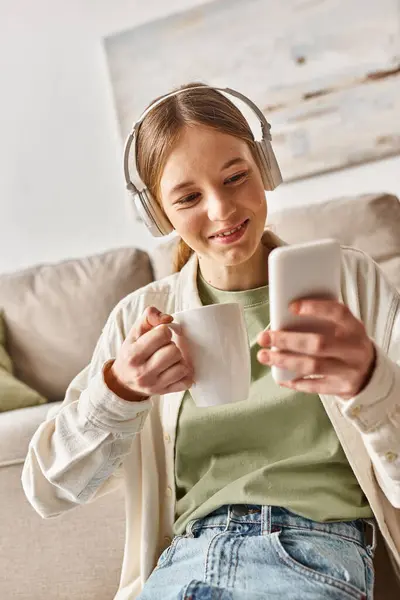 Carefree teenager girl in wireless headphones using smartphone while holding cup of tea at home — Stock Photo