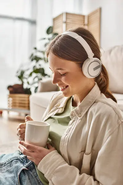 Unbekümmertes Teenager-Mädchen in ihren drahtlosen Kopfhörern, das Musik hört und zu Hause eine Tasse Tee hält — Stockfoto