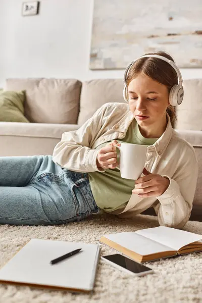 Relajada adolescente disfrutando de la música con auriculares y sosteniendo una taza cerca de los cuadernos en la alfombra - foto de stock