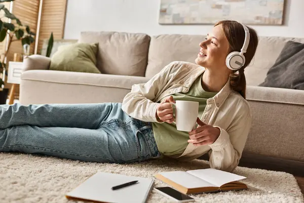 Relaxed gen z girl enjoying music with headphones and holding a mug near notebooks on carpet — Stock Photo