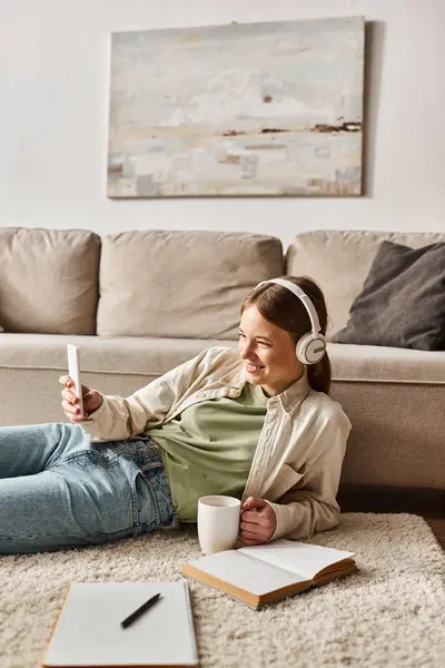 Entspanntes Gen-Z-Mädchen genießt Musik mit Kopfhörern und macht Selfie, während sie einen Becher in der Nähe von Notizbüchern hält — Stockfoto