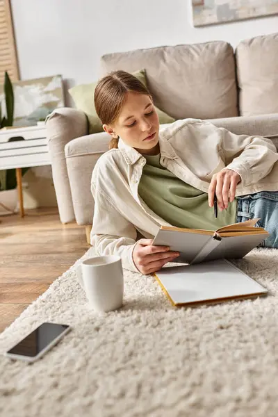 Adolescente niña leyendo libro mientras hace la tarea entre el dispositivo, y la taza de té en la alfombra, generación z - foto de stock