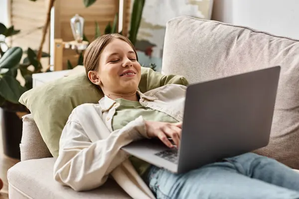 Adolescent gai utilisant son ordinateur portable et couché sur un canapé confortable à la maison, enseignement à distance — Photo de stock