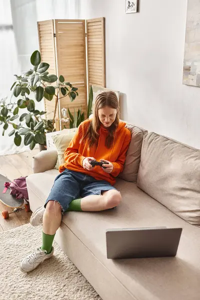 Emotionales Teenie-Mädchen, das mit Joystick und Laptop spielt, während es zu Hause auf dem Sofa sitzt, Wochenendstimmung — Stockfoto