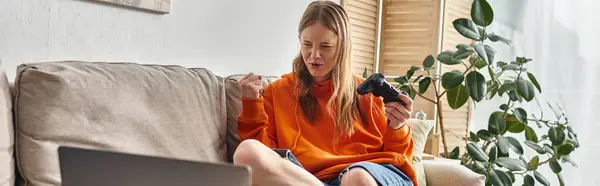 Emotional teen girl with joystick and laptop winning game and sitting on sofa at home, banner — Stock Photo