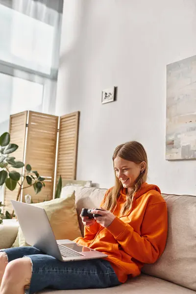Joyeuse adolescente jouant avec un joystick et un ordinateur portable tout en étant assis sur le canapé à la maison, vibes week-end — Photo de stock