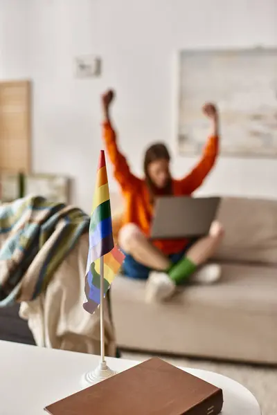 Fokus auf LGBTQ-Flagge auf Schreibtisch neben verschwommenem Teenager-Mädchen, das mit erhobenen Händen und Laptop sitzt — Stockfoto