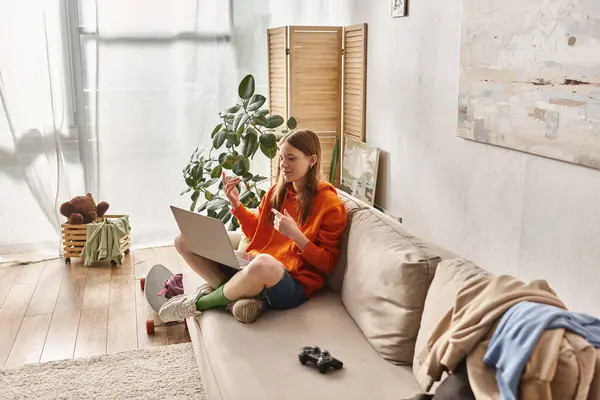 Счастливая девушка-подросток улыбается во время видеозвонка на ноутбуке и сидит рядом с джойстиком на диване — стоковое фото