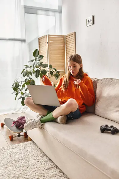 Gen z teenager girl smiling during a video call on a laptop and sitting near joystick on sofa — Stock Photo