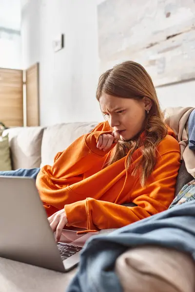 Adolescente enfocada ver una película en un ordenador portátil mientras se relaja en un sofá en la sala de estar, ocio - foto de stock