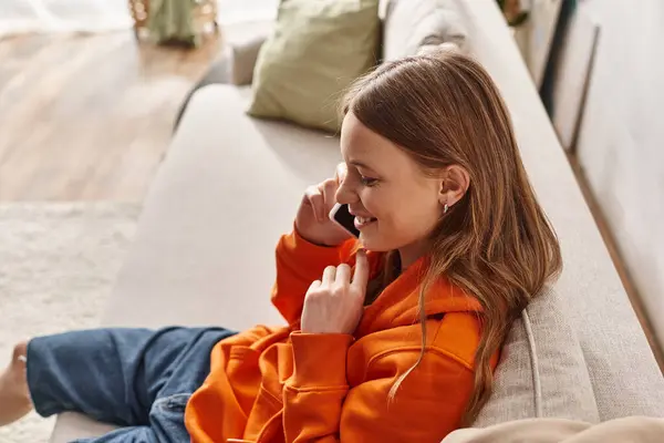 Щаслива дівчина-підліток, яка телефонує на смартфон і сидить на дивані у вітальні, гарний настрій — стокове фото