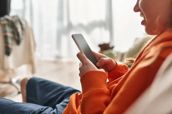 Cropped happy zoomer girl using her smartphone and sitting on sofa in living room, social media user — Stock Photo