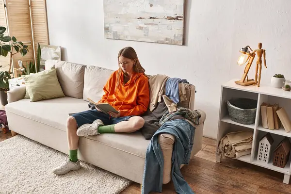 Adolescente leitura livro enquanto sentado no sofá bagunçado ao lado de pilha de roupas no apartamento moderno — Fotografia de Stock