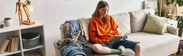 Teenage girl reading book while sitting on messy sofa next to pile of clothes in apartment, banner — Stock Photo