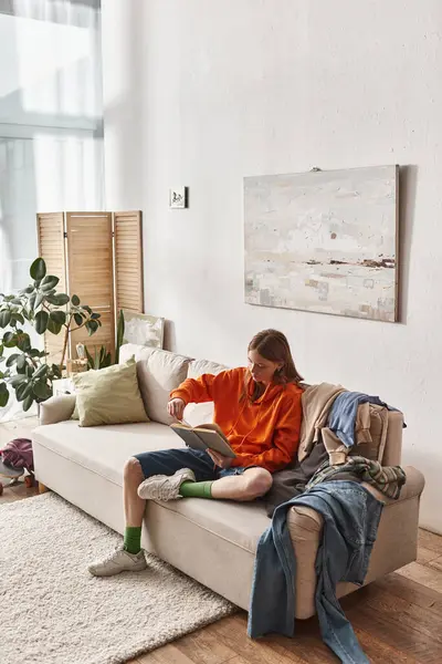 Inteligente adolescente leyendo libro mientras está sentado en sofá desordenado al lado de la pila de ropa en el apartamento - foto de stock