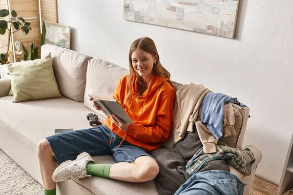 Glücklicher Teenager liest Buch, während er auf dem Sofa neben chaotischem Kleiderstapel in der Wohnung sitzt — Stockfoto