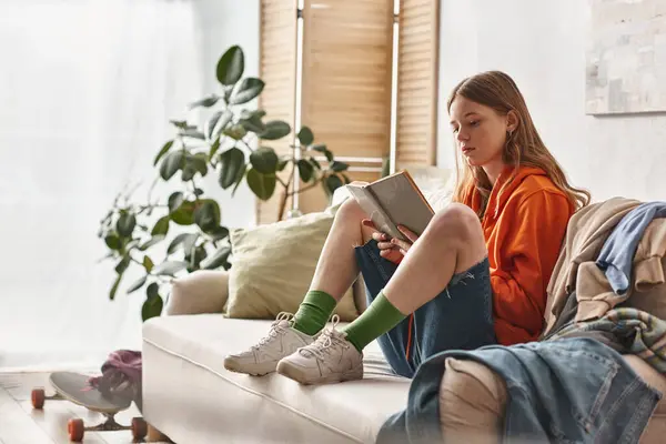Adolescente dedicado a la lectura de libros y sentado en el sofá junto a la pila desordenada de ropa en el apartamento - foto de stock