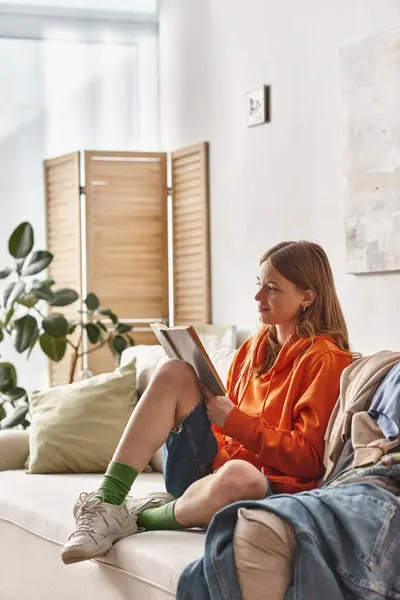 Dreamy teenage girl engaged in reading book and sitting on sofa next to messy pile of clothes — Stock Photo