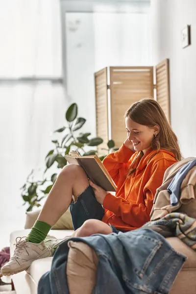 Adolescent souriant engagé dans la lecture du livre et assis sur le canapé à côté de tas de vêtements désordonnés — Photo de stock