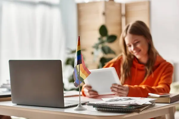 Cheerful teenage girl with tablet engaging in e-learning process near lgbtq pride flag on desk — Stock Photo