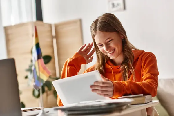 Cheerful teenage girl with tablet saluting during e-learning process near lgbtq pride flag on desk — Stock Photo