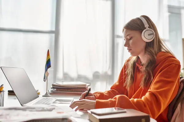 Focused young teenage girl in wireless headphones e-learning beside pride flag and stationery — Stock Photo